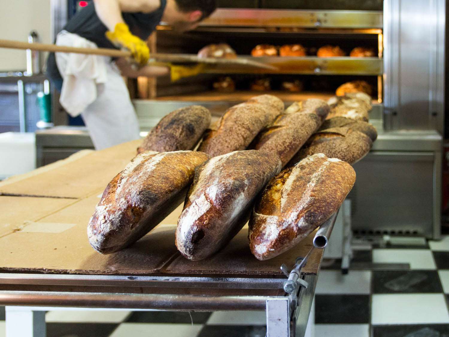 Loaves of bread on a baker's table in the kitchen at Tartine Bakery. In the background, someone is using a long handled peel to move loaves of bread in a commercial oven. 