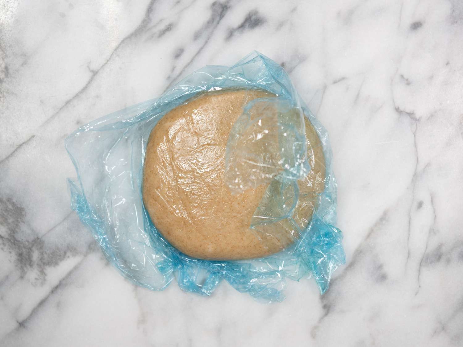 The dough, half-wrapped in plastic and placed on a white marble slab.