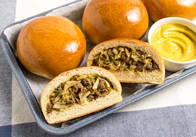 3 Bierocks on a platter with small white mustard dish, with a 4th bun cut open and filling is visible. Platter is on a grey white and blue striped textile. 