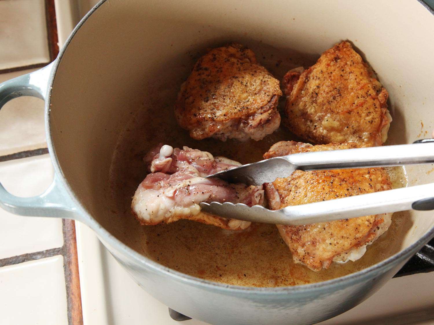 The browned chicken thighs are turned over with a pair of tongs to cook on the other side.
