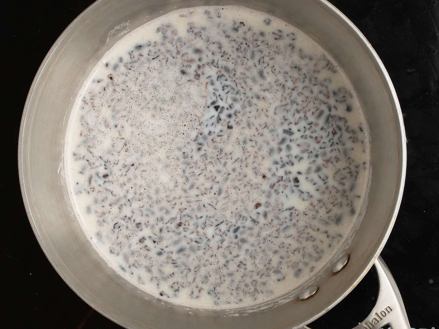 A stainless steel saucepan holding cocoa nibs stirred into simmering milk.