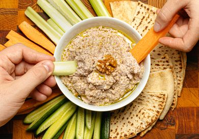 Two hands holding slices of vegetable dipping into a bowl of walnut skordalia surrounded by flatbread and vegetable sticks