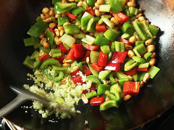 Adding minced garlic, ginger, and scallions to stir-frying peppers, celery, and peanuts in a wok for takeout-style kung pao chicken.