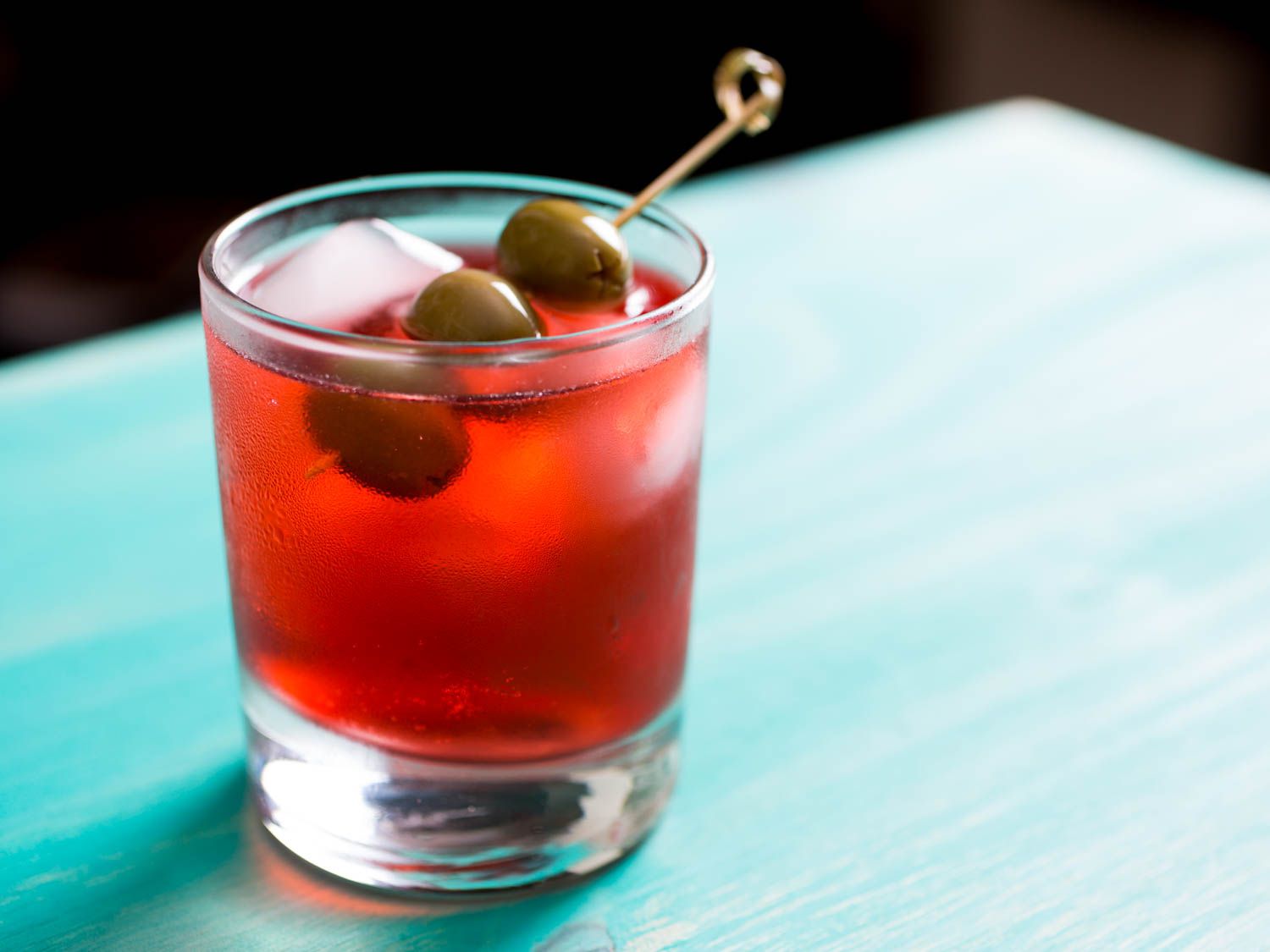 A Campari Spritz in a rocks glass, garnished with 3 olives on a pick. 