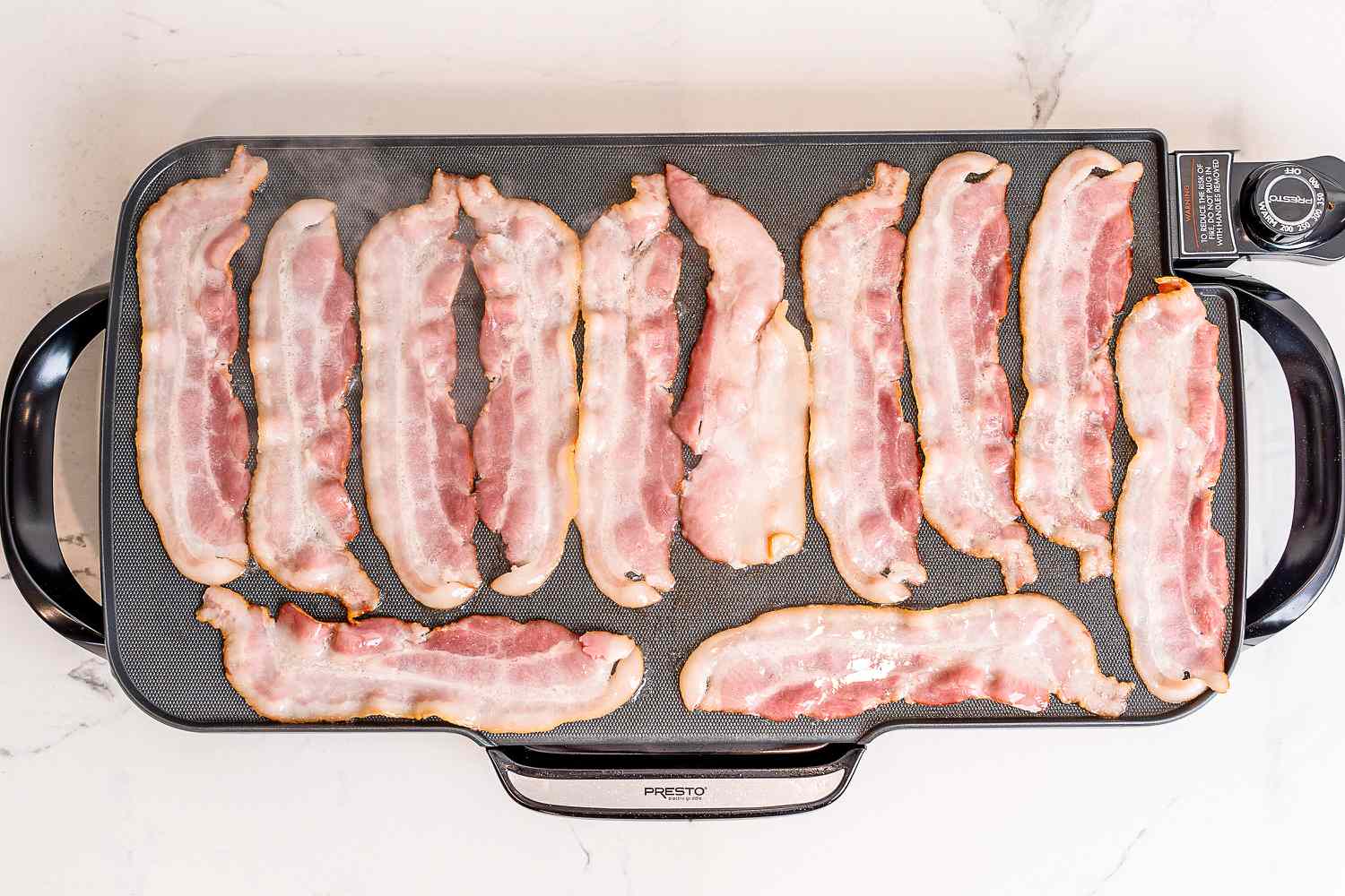 Several slices of bacon cooking on an electric griddle