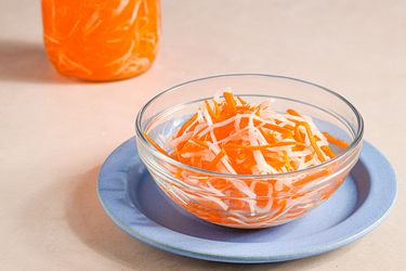 A small glass bowl on a blue plate holding Vietnamese pickled carrots and daikon. There is a jar of pickles and bring in the top lefthand corner of the image.