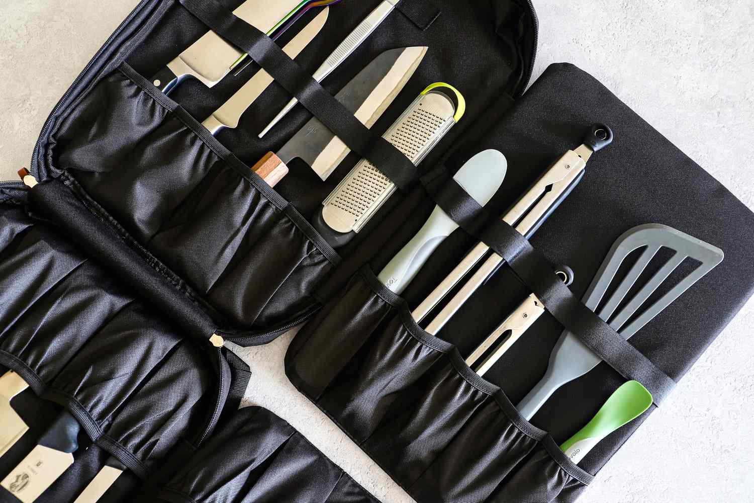 A black carrying case containing various kitchen tools including knives a grater tongs and spatulas organized in pockets