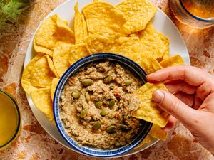 A hand dipping a tortilla chip into a bowl of dip garnished with seeds surrounded by tortilla chips on a plate