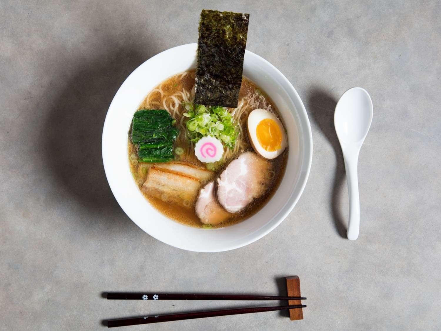 Overhead shot of a bowl of shoyu ramen, garnished with meats, seaweeds, fish cake, and egg, next to a spoon and a pair of chopsticks