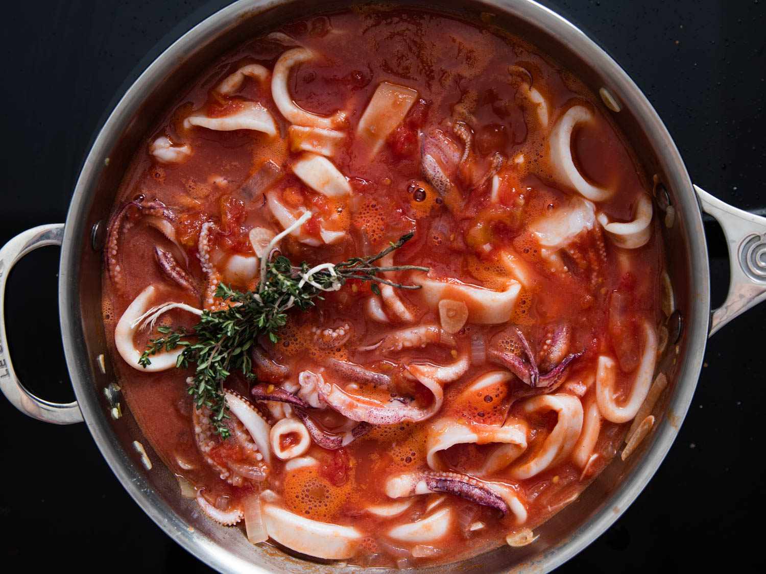 Overehead view of squid, onions, garlic, crushed tomatoes, and bundled thyme sprigs simmering in a saucepan.