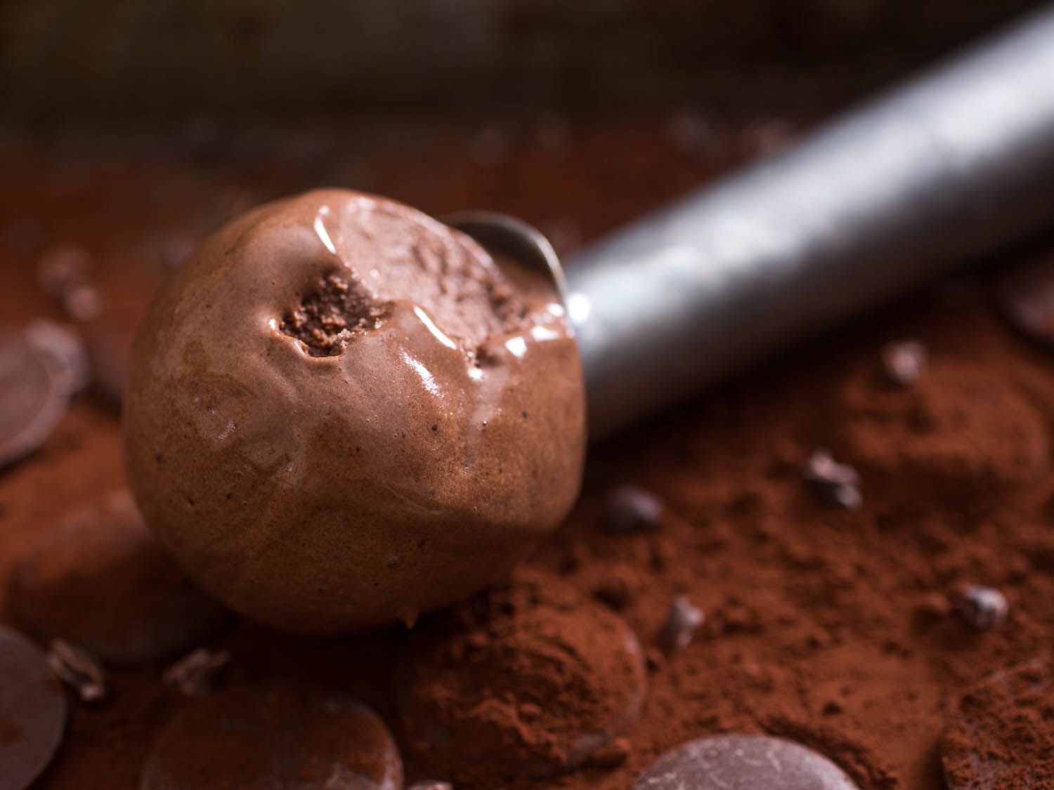 A scoop of darkest dark chocolate ice cream with cocoa powder, chocolate chips, and chocolate disks in the background.