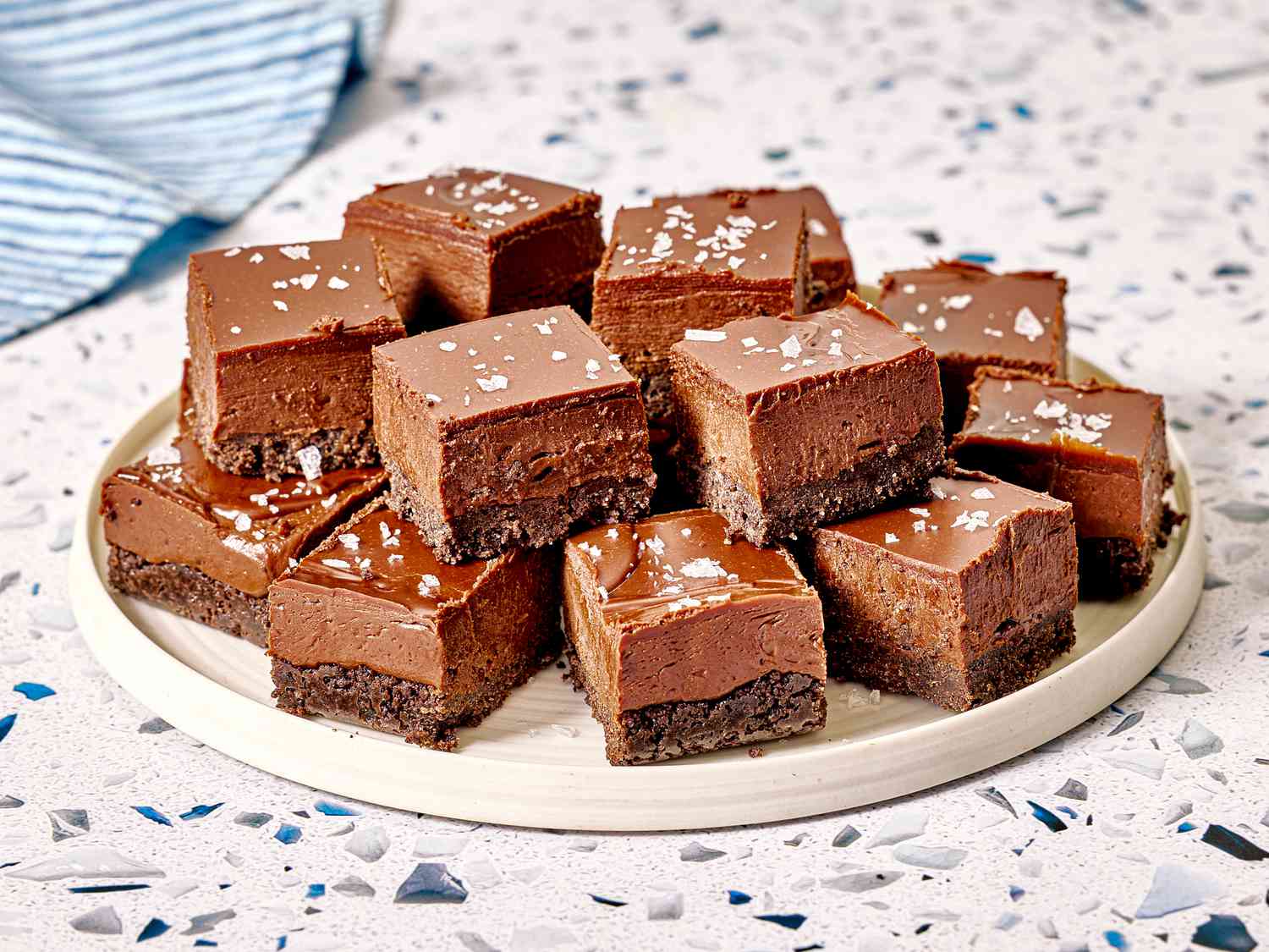 A plate of layered chocolate squares with a fudgy and textured appearance topped with sea salt