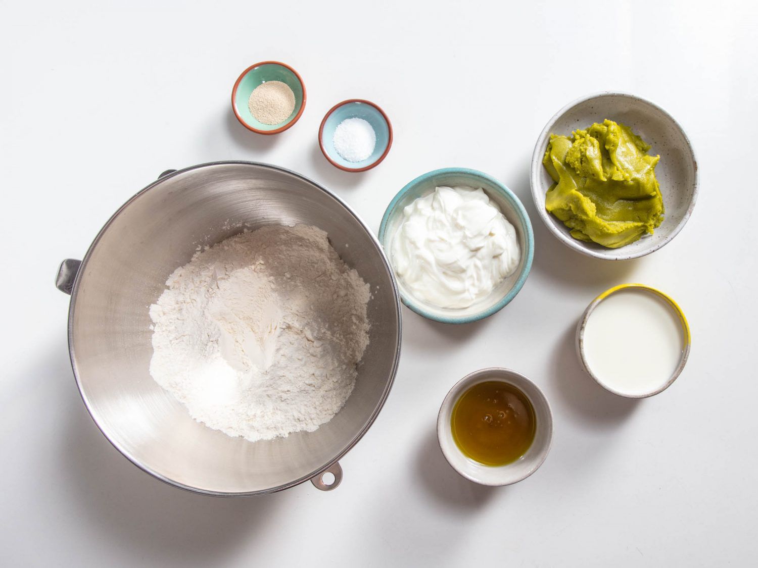 Averhead shot of little bowls containing flour, salt, yeast, Greek yogurt, pistachio oil, pistachio paste, and milk