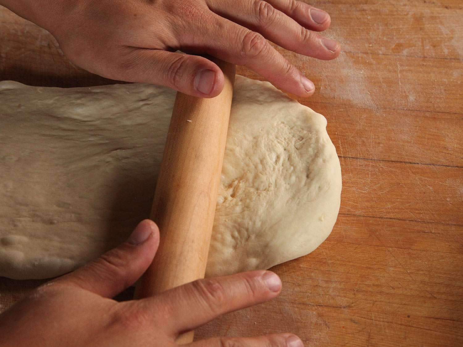 A pair of hands using a rolling pin to roll out dough on a wooden surface