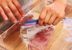 Two hands placing a Ziplock bag with meat into it into a tub filled with water. This image displays how to use the "displacement method" for removing air from a bag without a vacuum sealer.