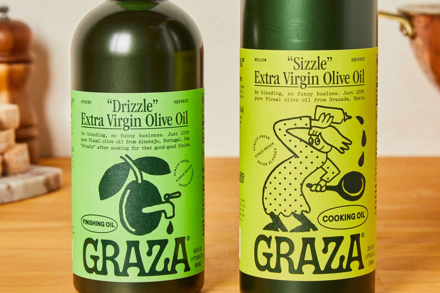 Two bottles of Graza Extra Virgin Olive Oil labeled Drizzle and Sizzle on a wooden table surface