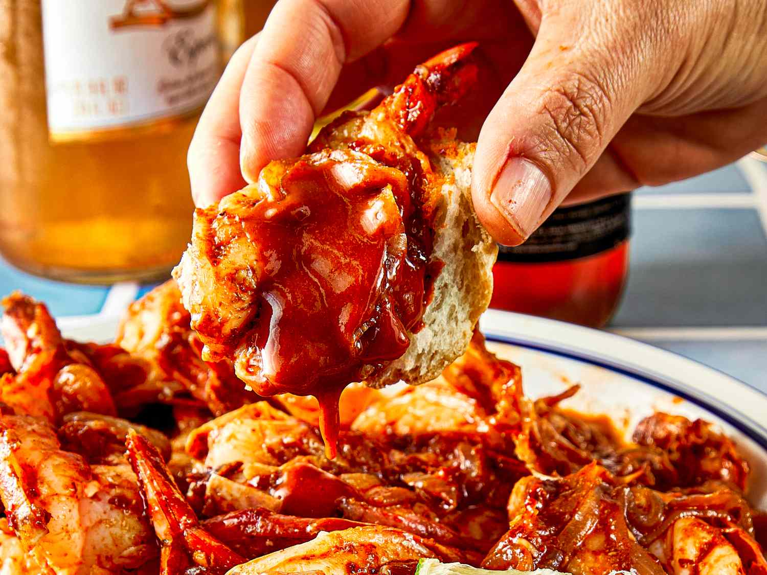 A hand holding a piece of food dipped in a spicy sauce with a plate of similar food and bottles in the background
