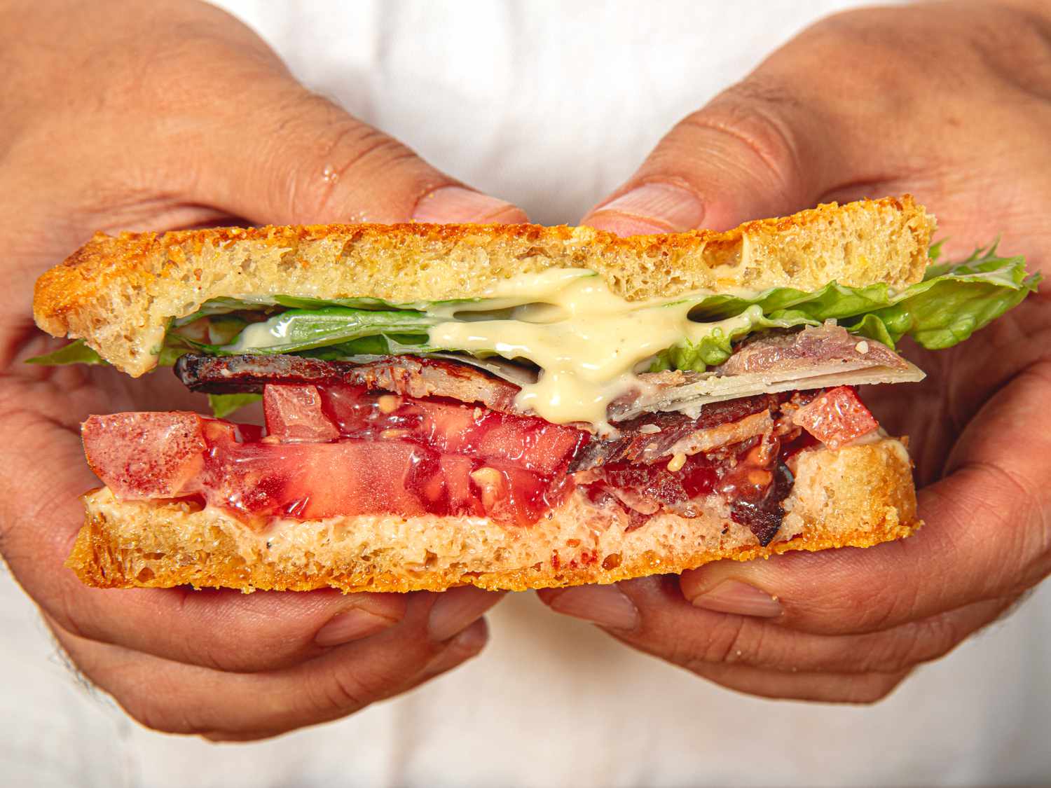 Two hands holding half of this the Caesar BLT sandwich, dripping with Caesar mayo