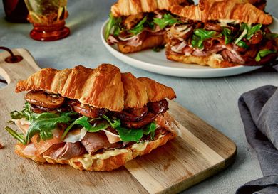 A sandwich made with puff pastry filled with beef greens and toppings served on a cutting board other sandwiches visible in the background
