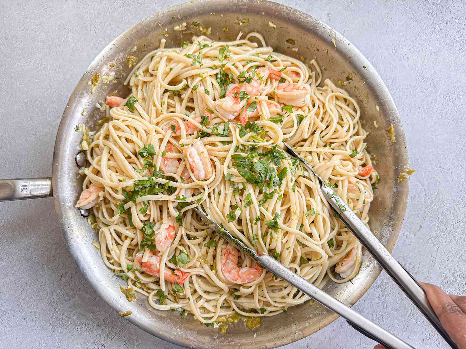 Shrimp and pasta dish in a skillet, garnished with chopped herbs, with tongs being used