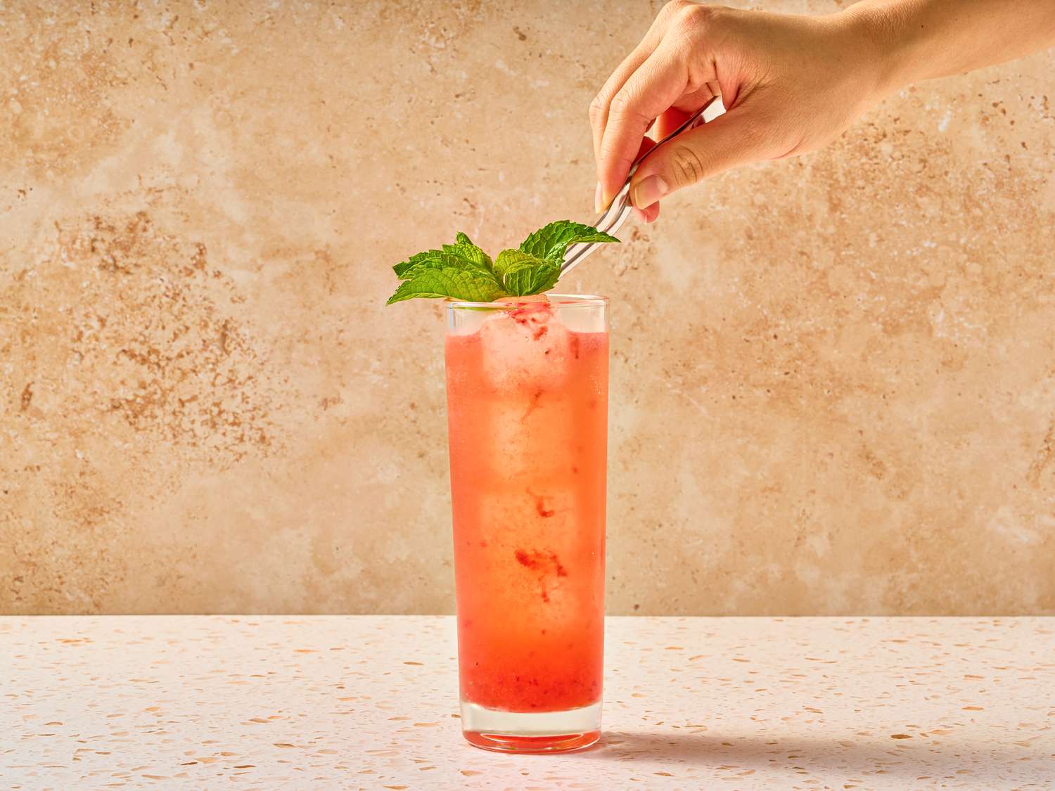 A tall glass with a pink beverage and mint leaf garnish a hand adjusts the garnish