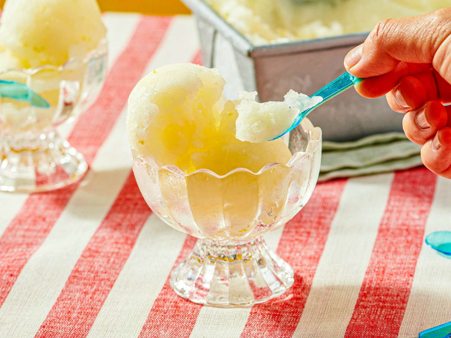 A hand scooping lemon sorbet from a glass bowl with a blue spoon placed on a red and white striped tablecloth