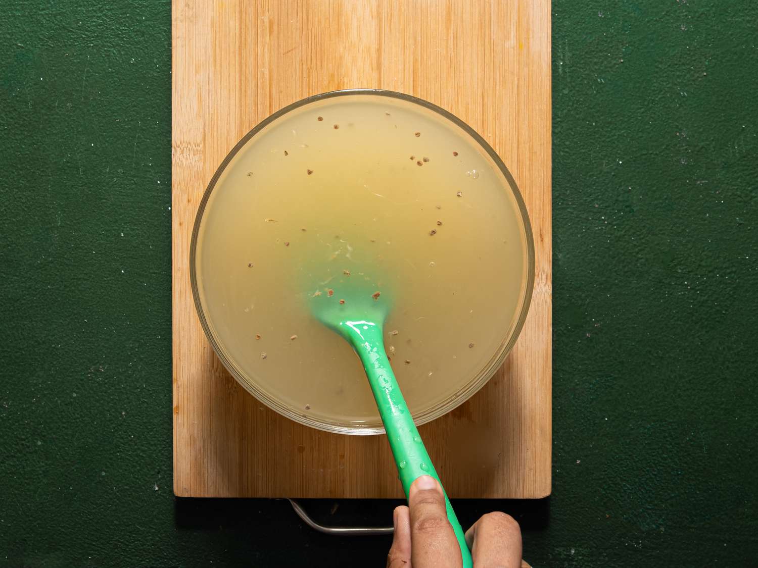 A person stirring a bowl of liquid with a green spoon on a wooden board