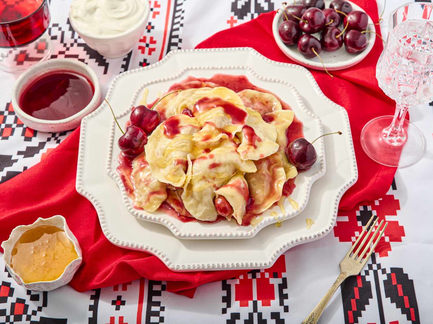 Cherry verenyky on decorative plates, on a printed surface, with a red napkin, and sides of cherry sauce, cream, and honey 