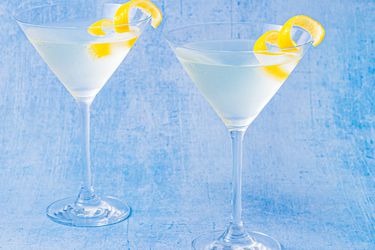 Two Martinis served with lemon twists on a distressed light blue surface.