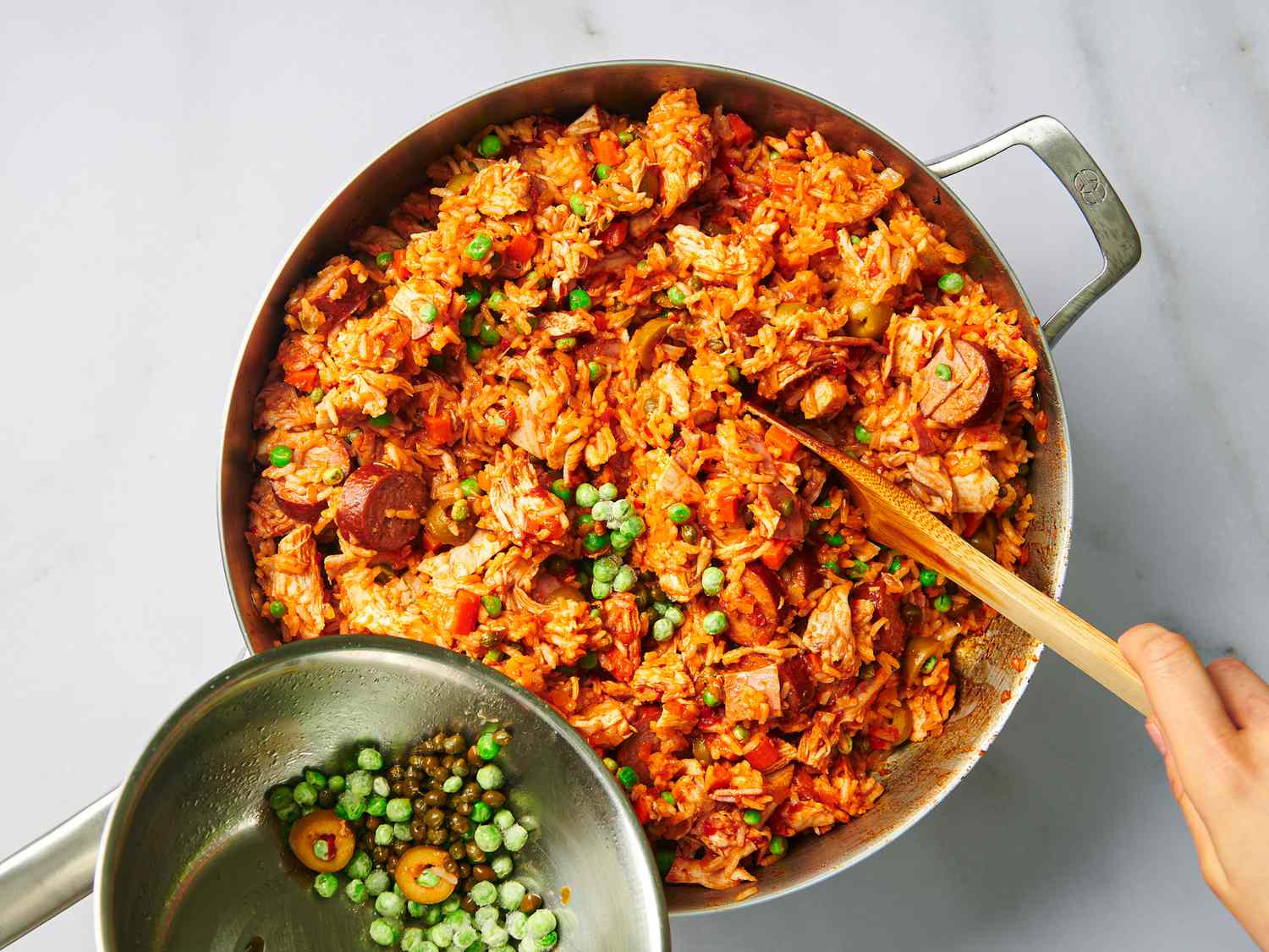 Capers, olive, and peas being stirred into the contents of the stainless steel pan, which now holds both the cooked rice and the chicken mixture.
