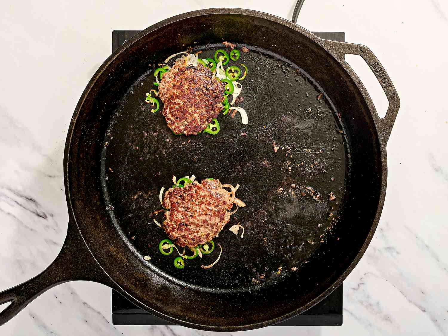 Two patties cooking in a skillet with sliced green chilies and onions