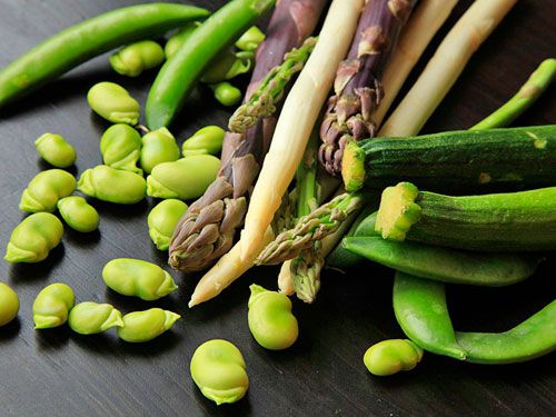 Fava beans, asparagus, snap peas, and zucchini on a black surface.