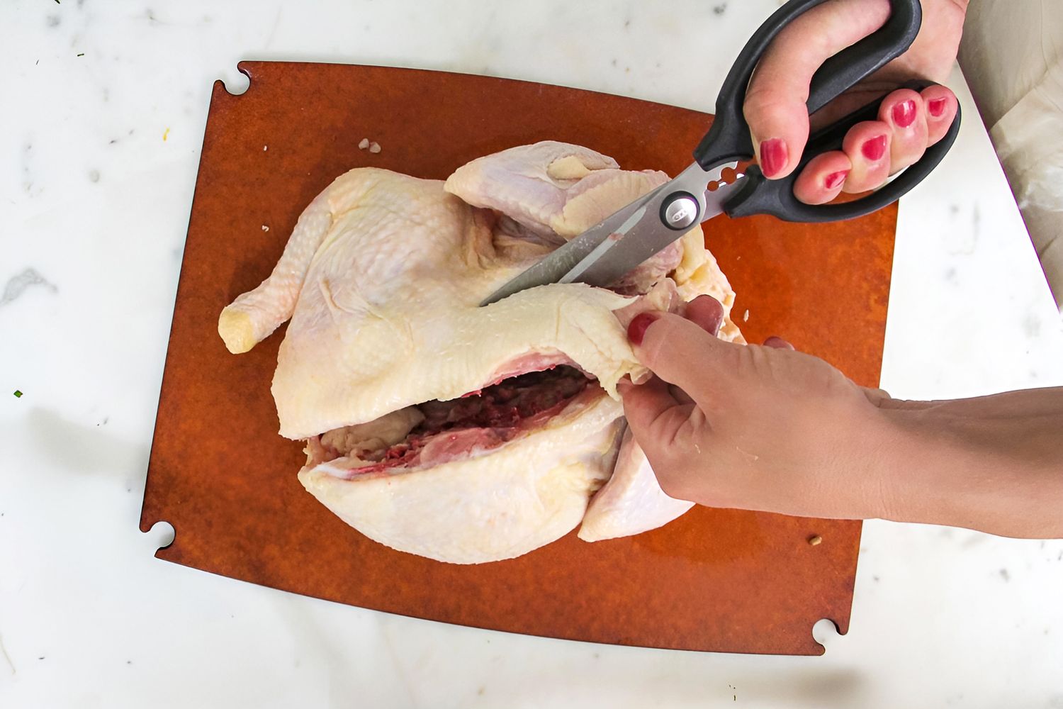 A person using kitchen scissors to cut through a raw chicken on a cutting board