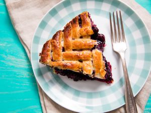 A wedge of herringbone lattice–topped blueberry pie on a plaid-rimmed plate with a fork