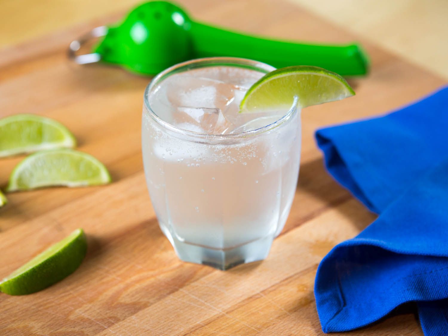 A gin rickey in a highball glass with a lime wedge on the rim. The glass is on a wood cutting board with a blue napkin, three other lime wedges, and a green hinged citrus juicer. 