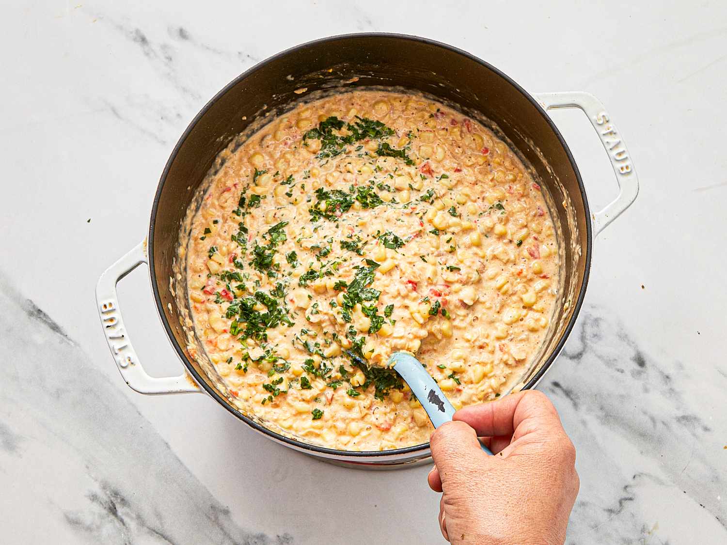 A hand stirring creamy corn in a pot with herbs on top
