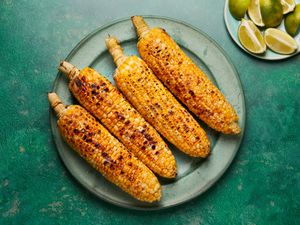 Four grilled corn cobs on a plate, accompanied by lime wedges on the side