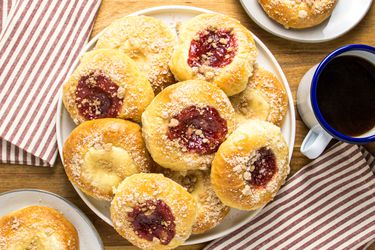 Kolaches on a platter, with one cream and one strawberry on 2 plates, with coffee on the side, striped napkins, on a wooden surface. 