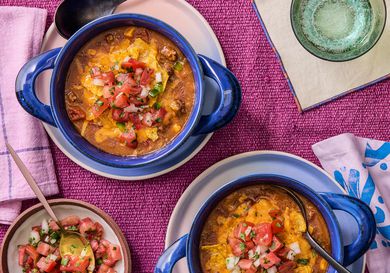 Overhead of 2 blue bowls with taco soup, on with a spoon in it. A small dish with pico de gallo on it. Pink napkins and sparkling water and a magenta colored textile surface. 