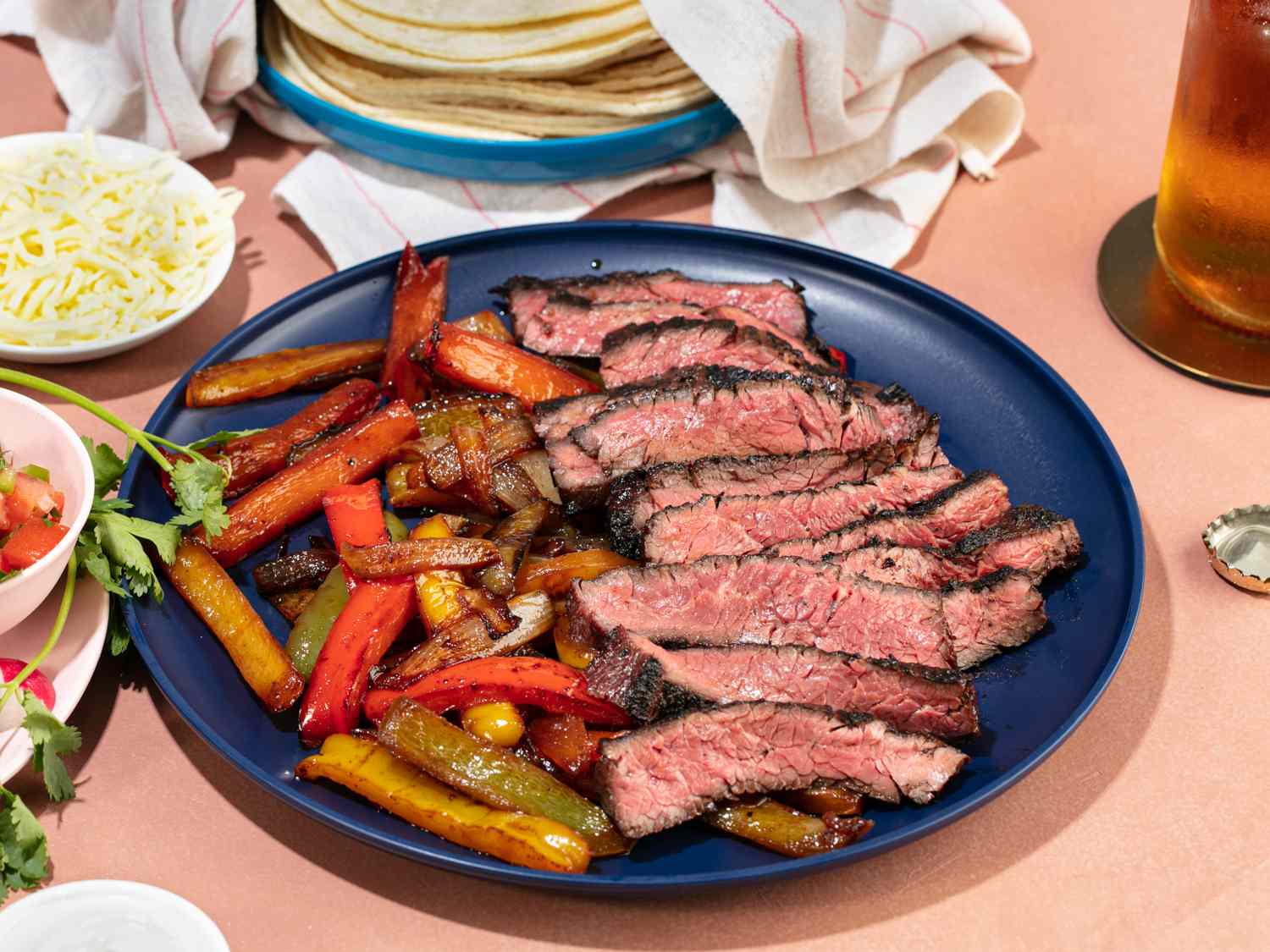 Grilled skirt steak fajitas cut into slices and grilled vegetables on a blue plate, next to a plate of tacos and a few bowls of condiments. 
