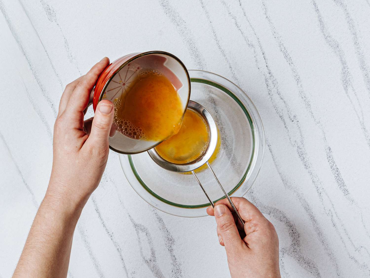 pouring mixture through a fine-mesh sieve into a glass bowl, on a white marbled surface 