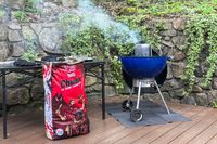 A barbecue setup with a bag of Jealous Devil Chunx Lump Charcoal and a grill chimney starter emitting smoke on a wooden deck