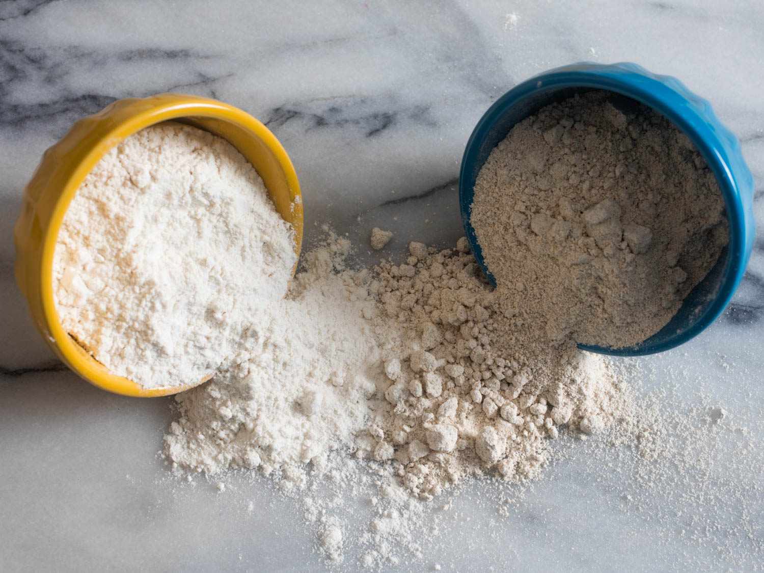 Yellow bowl of all-purpose flour and blue bowl of oat flour tipping out onto a marble countertop.