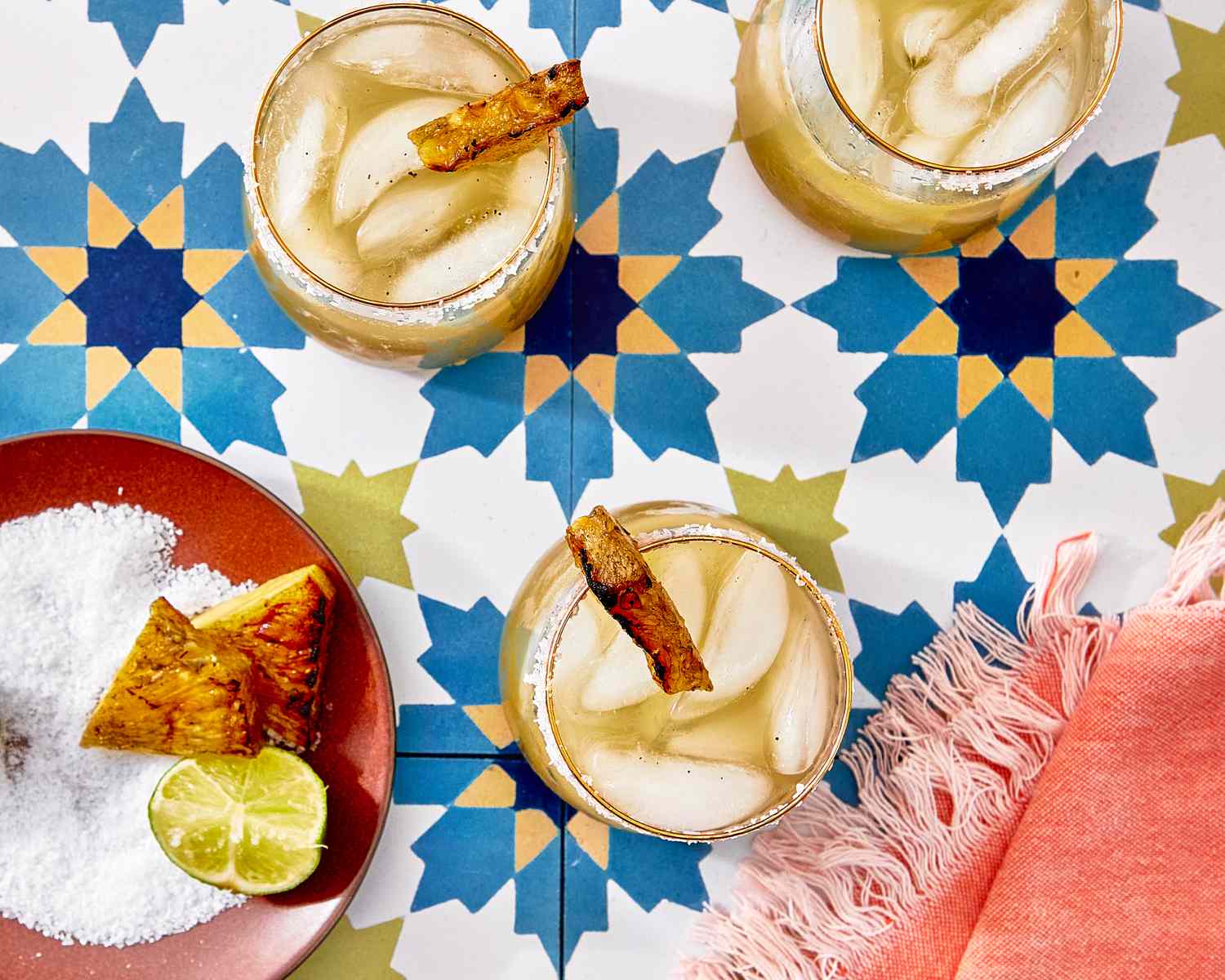 Three margarita glasses garnished with grilled pineapple on a patterned background, with lime wedges and a salt dish nearby