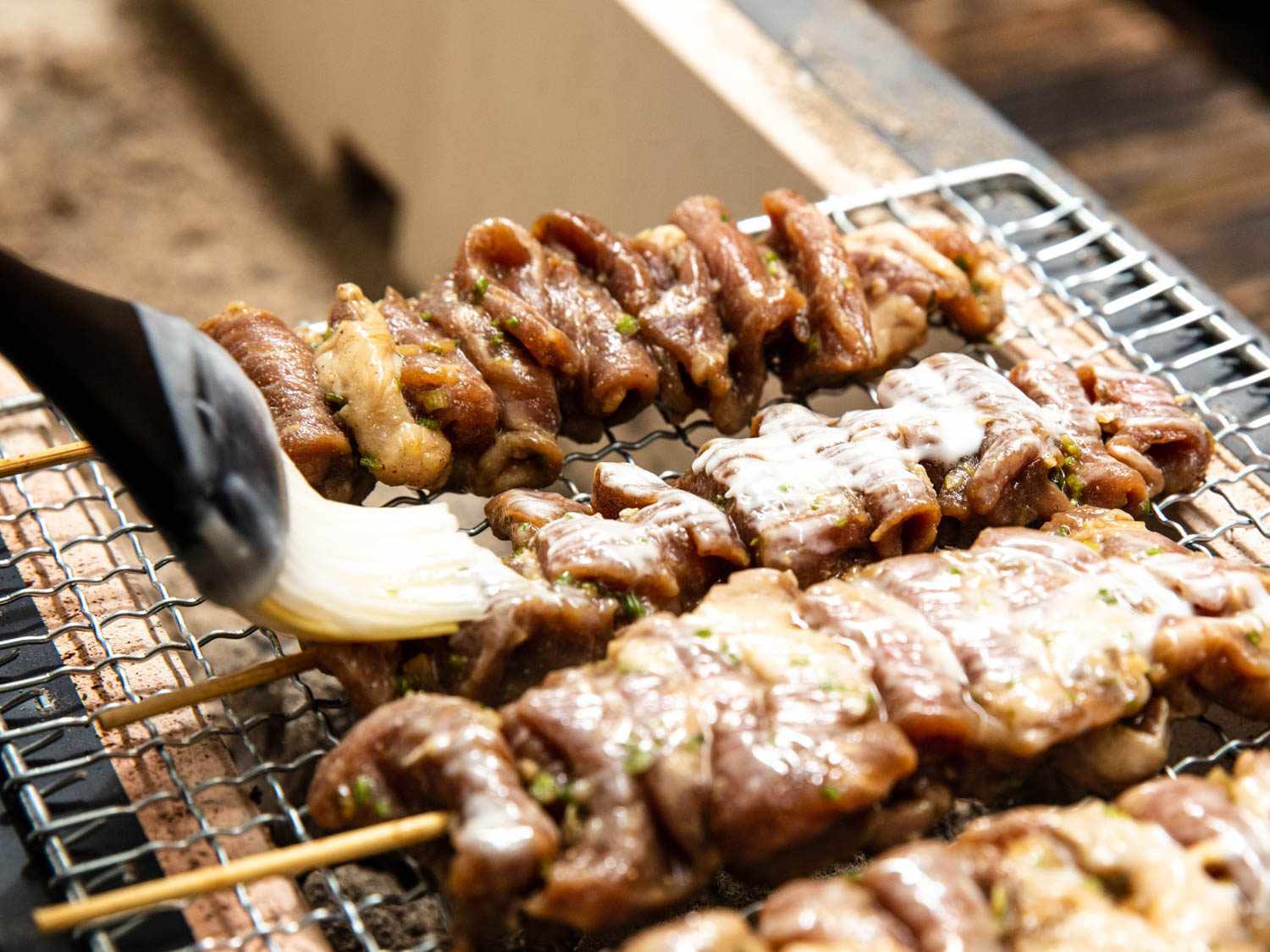 Close-up of Thai pork skewers being brushed with a pastry brush while resting on a wire mesh rack, suspended over coals