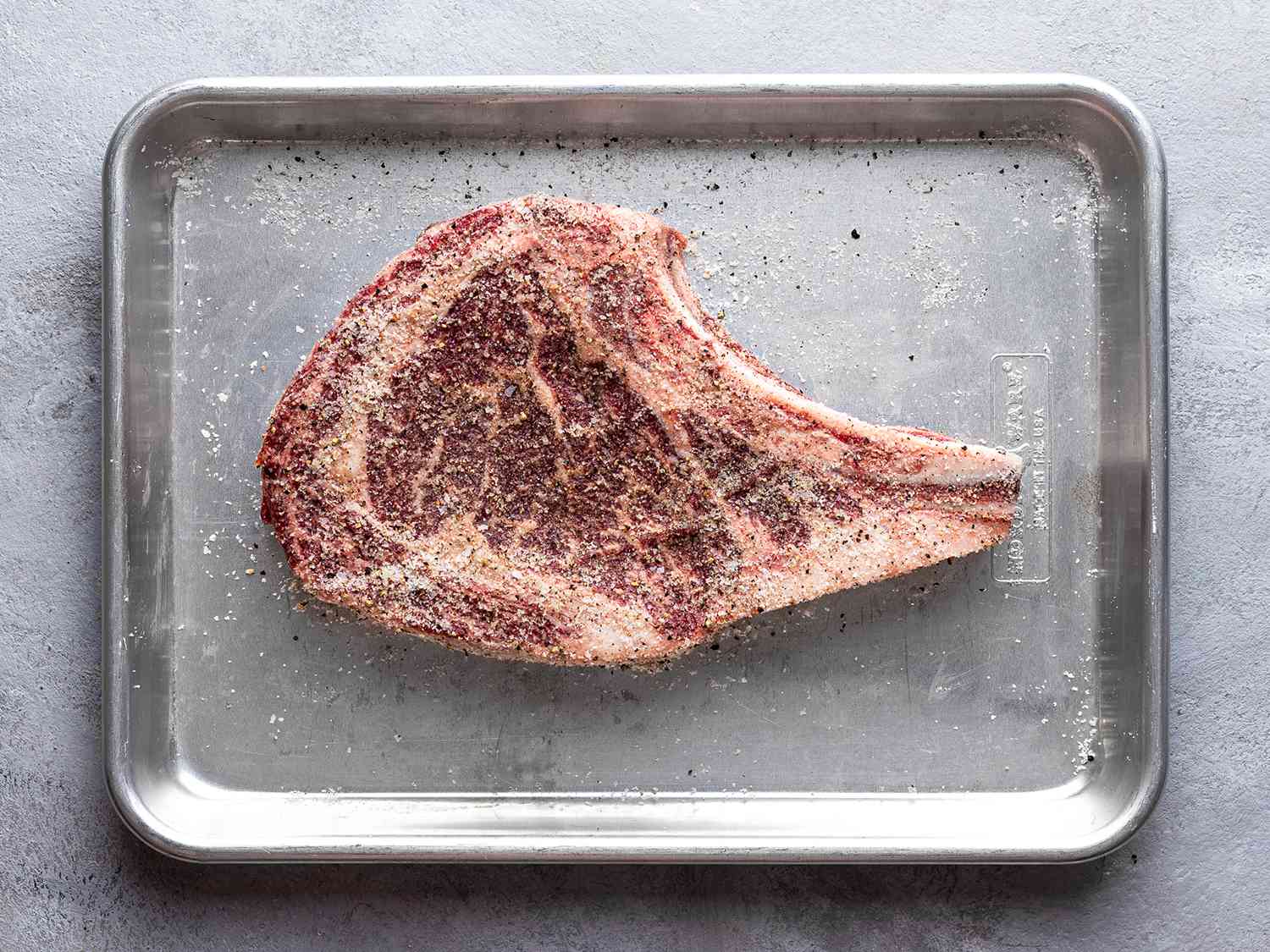 A salted bone-in ribeye steak on a quarter sheet pan.