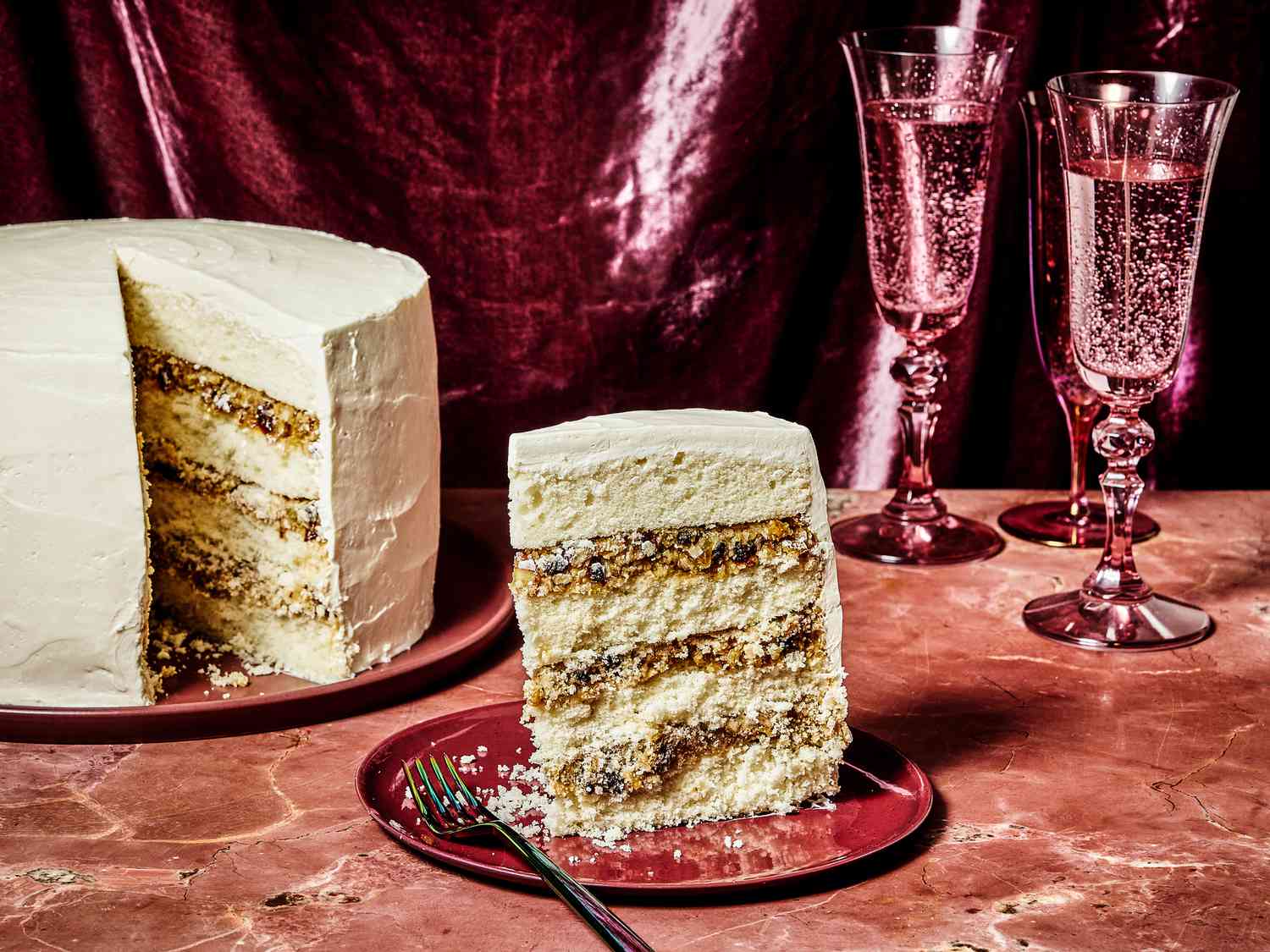 Alabama lane cake, with a slice placed on a red plate, on a red marble tabletop. 2 Magenta glasses of bubbly water, and a magenta fabric draped in the background. 