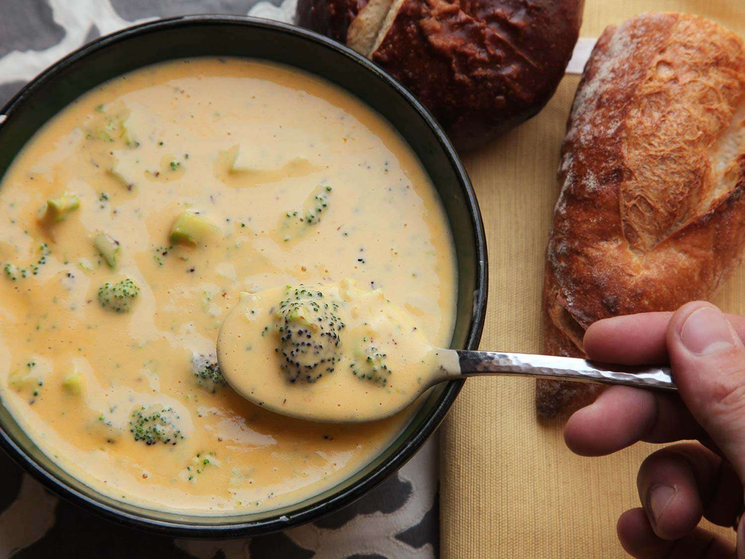 A spoon scooping up broccoli cheese soup in a bowl, next to half a loaf of crusty bread. 
