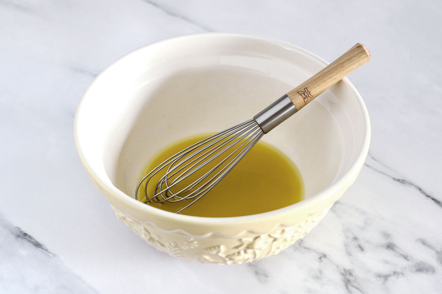 A whisk resting in a Mason Cash bowl containing a yellow liquid on a table surface