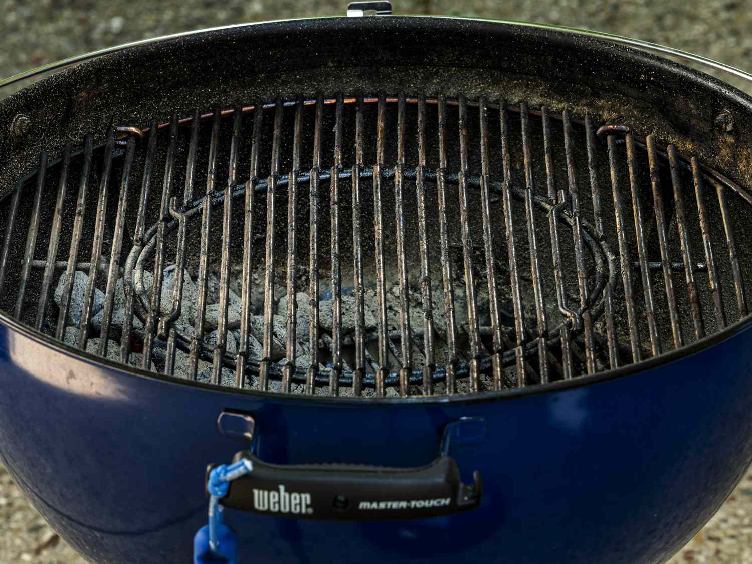 A blue Weber-brand charcoal grill, showing that the coals are spread to one side of the interior of the basin.
